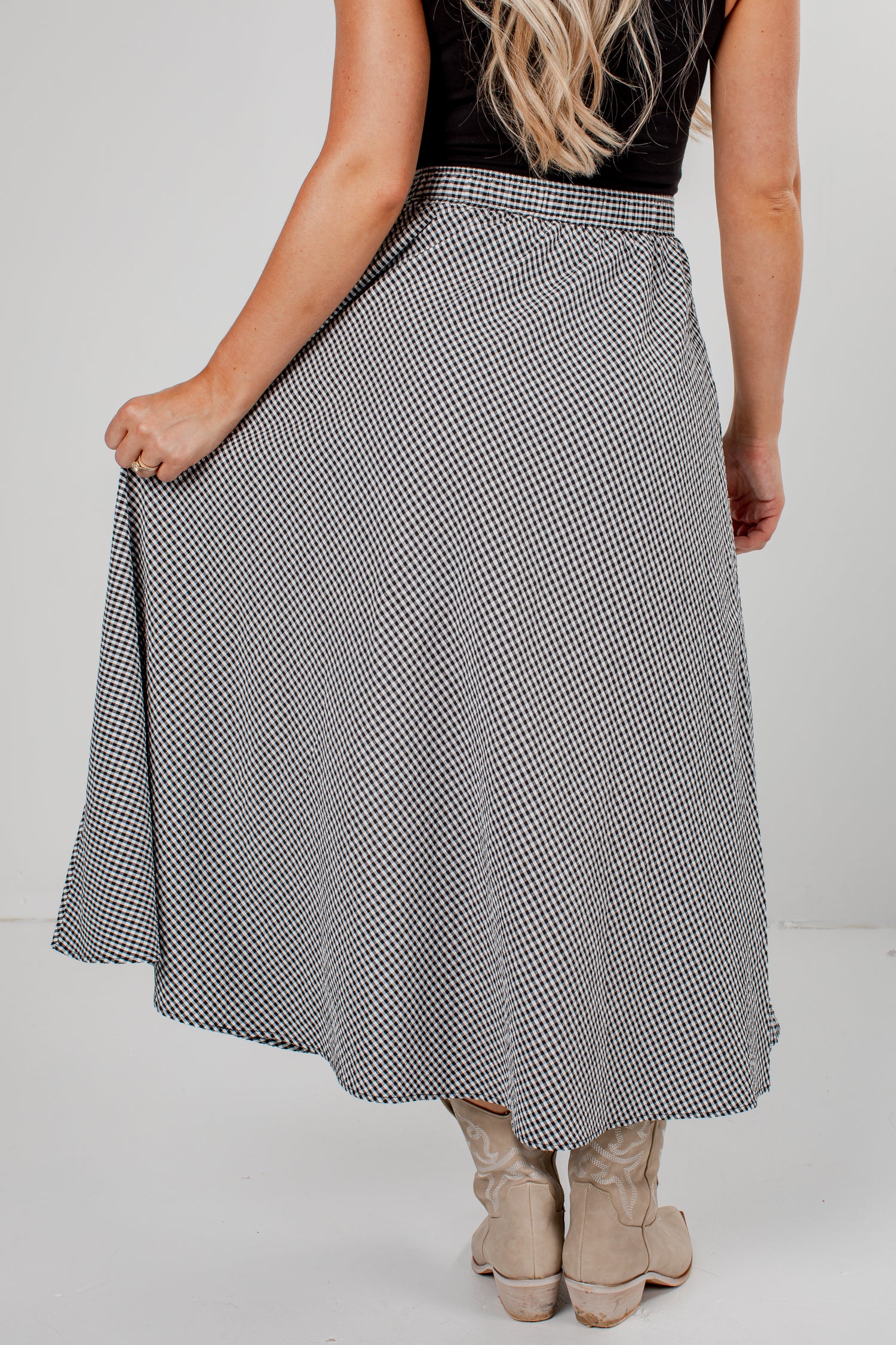A woman wearing a black and white micro-checkered midi skirt, paired with a black sleeveless top and beige cowboy boots, shown from the back against a white studio background.