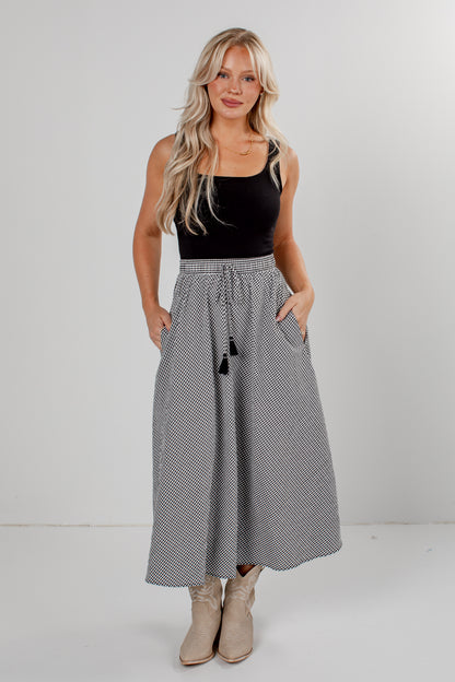 A woman modeling a high-waisted, maxi-length skirt with a black and white micro-check pattern, a drawstring waist, and black tassels, paired with a black sleeveless top and beige boots.