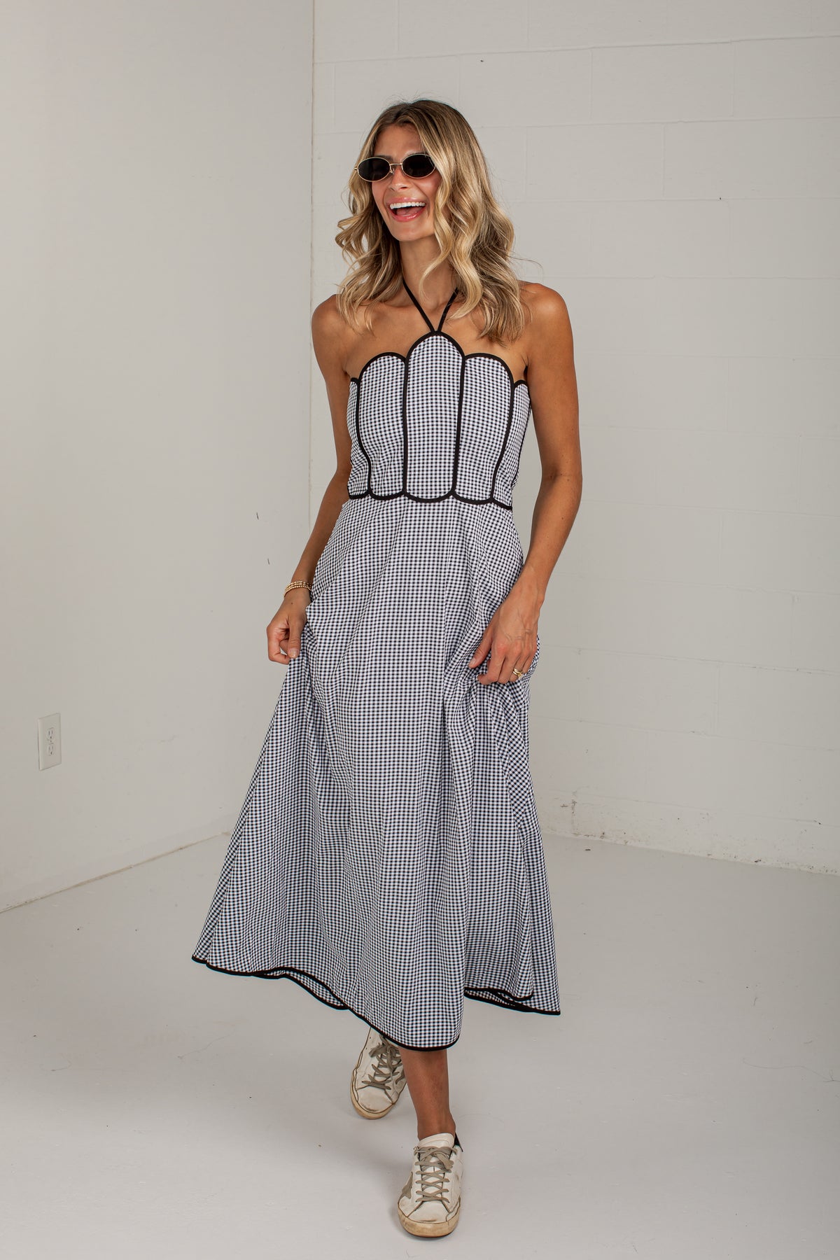 A woman modeling a sleeveless midi dress with thin vertical black and white stripes, featuring a scalloped corset-style bodice and a flared skirt, paired with beige sneakers against a white studio background.