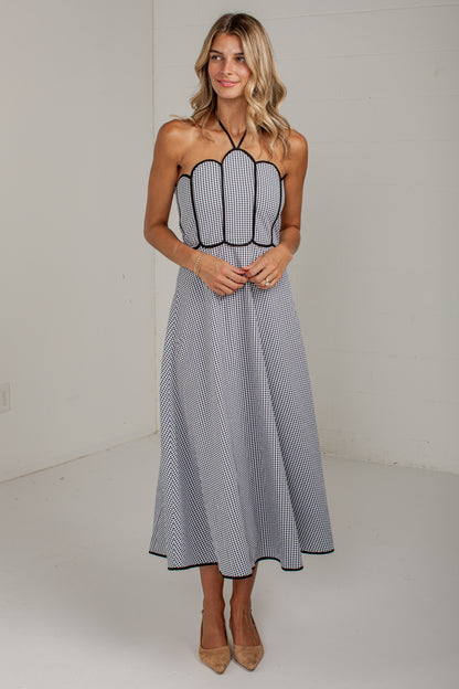 A woman modeling a sleeveless halter midi dress with a black and white gingham pattern, black piping, and a flared skirt, standing against a plain white studio background.