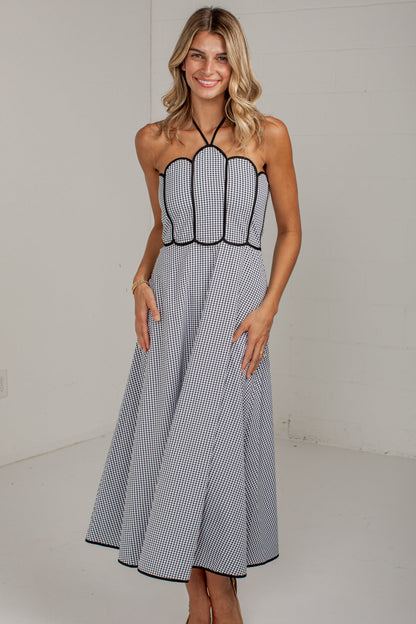 A woman modeling a sleeveless halter-neck midi dress with a black and white gingham pattern and black trim, standing against a plain white studio wall.