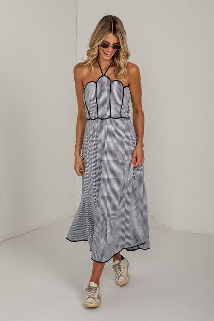 A woman modeling a sleeveless halter-neck maxi dress with a black and white pinstripe pattern, featuring a scalloped bodice and a flowing skirt, paired with beige sneakers in a minimalist white studio setting.