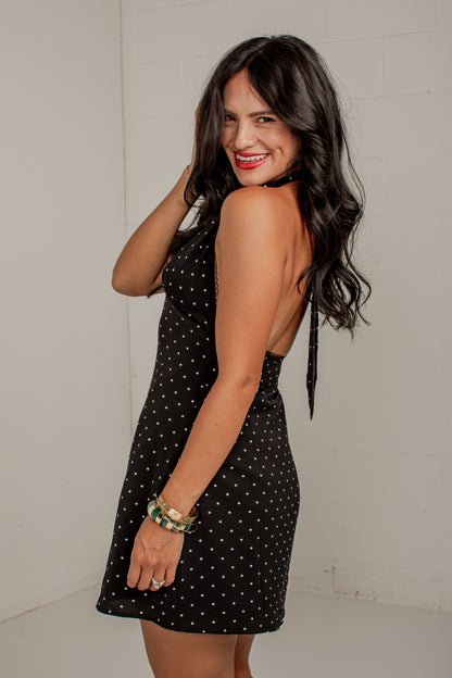 A woman modeling a black sleeveless mini dress with white polka dots, featuring a backless open back and tie detail at the neck, set against a plain light gray wall.