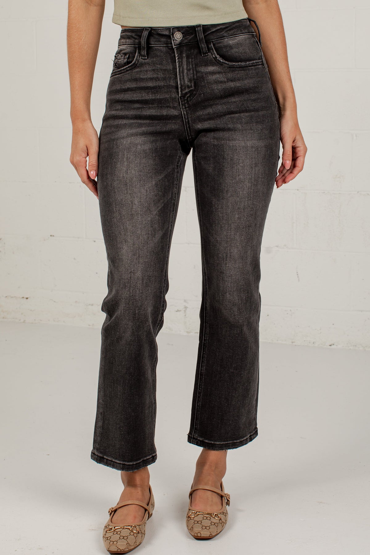 A model wearing a pair of black washed denim jeans with a straight-to-crew leg, paired with a light beige top and brown flat shoes, standing against a white brick wall.