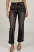 A model wearing a pair of black washed denim jeans with a straight-to-crew leg, paired with a light beige top and brown flat shoes, standing against a white brick wall.