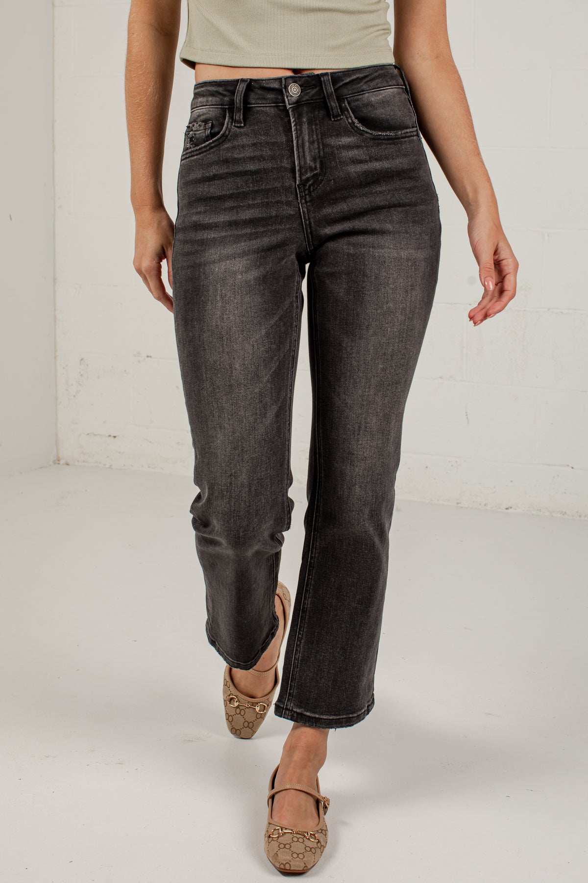 A model wearing a pair of dark gray washed straight-leg jeans, styled with a light beige crop top and brown flat shoes, standing against a white studio wall.