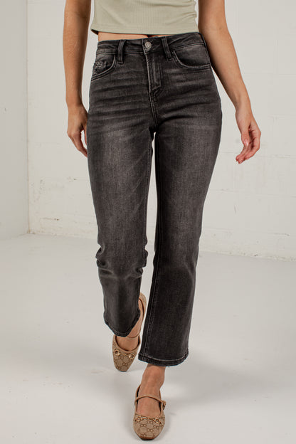 A model wearing a pair of dark gray washed straight-leg jeans, styled with a light beige crop top and brown flat shoes, standing against a white studio wall.