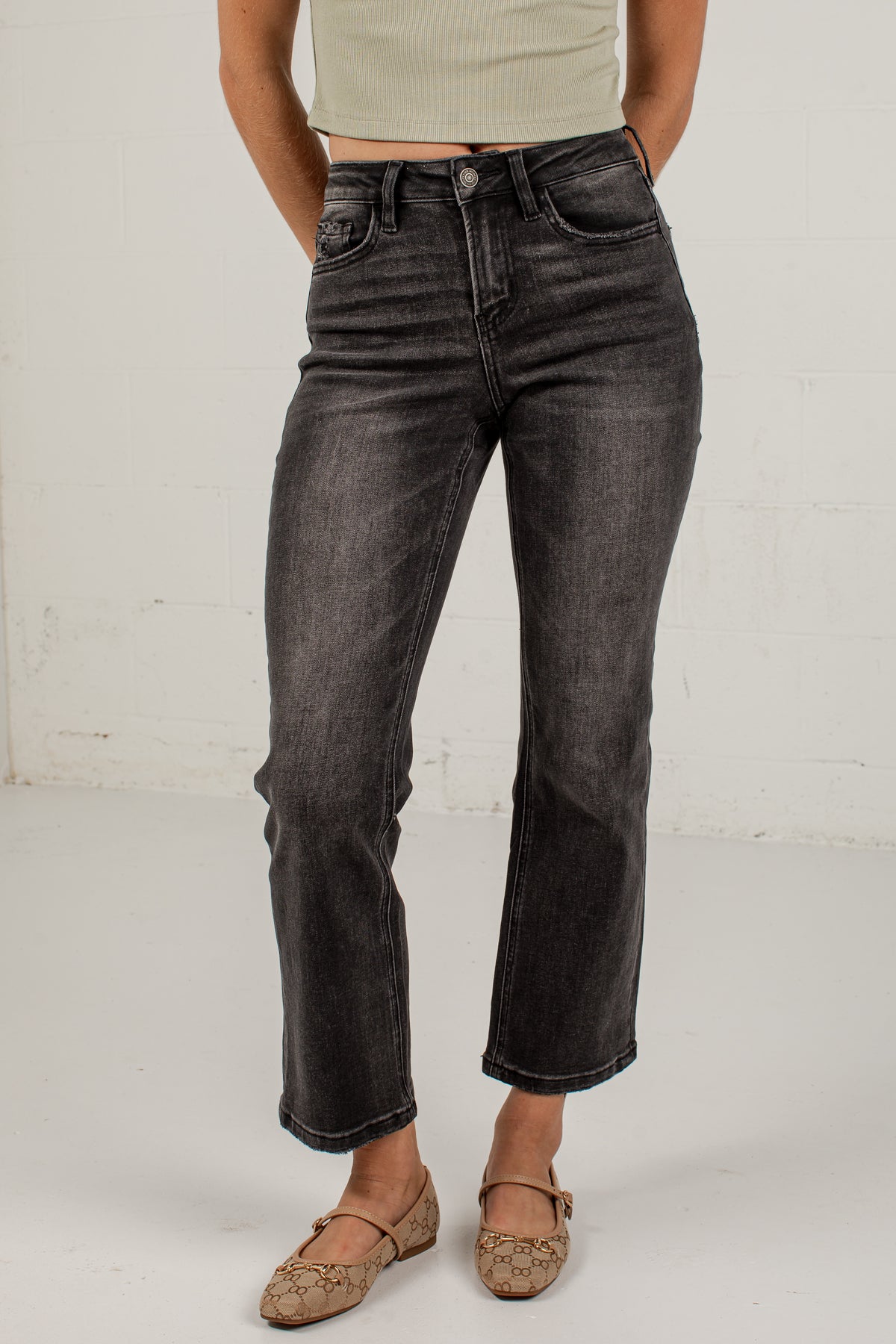 A person wearing dark gray washed cropped jeans with a straight-to-medium leg, paired with a light beige ribbed crop top and brown flat shoes, standing against a white brick wall.