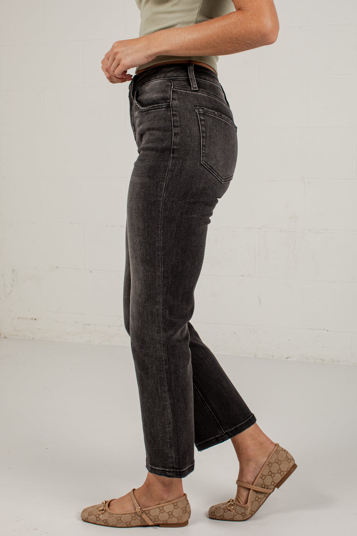 A person shown from the waist down wearing dark gray washed cropped jeans and beige flat shoes, standing against a white wall.