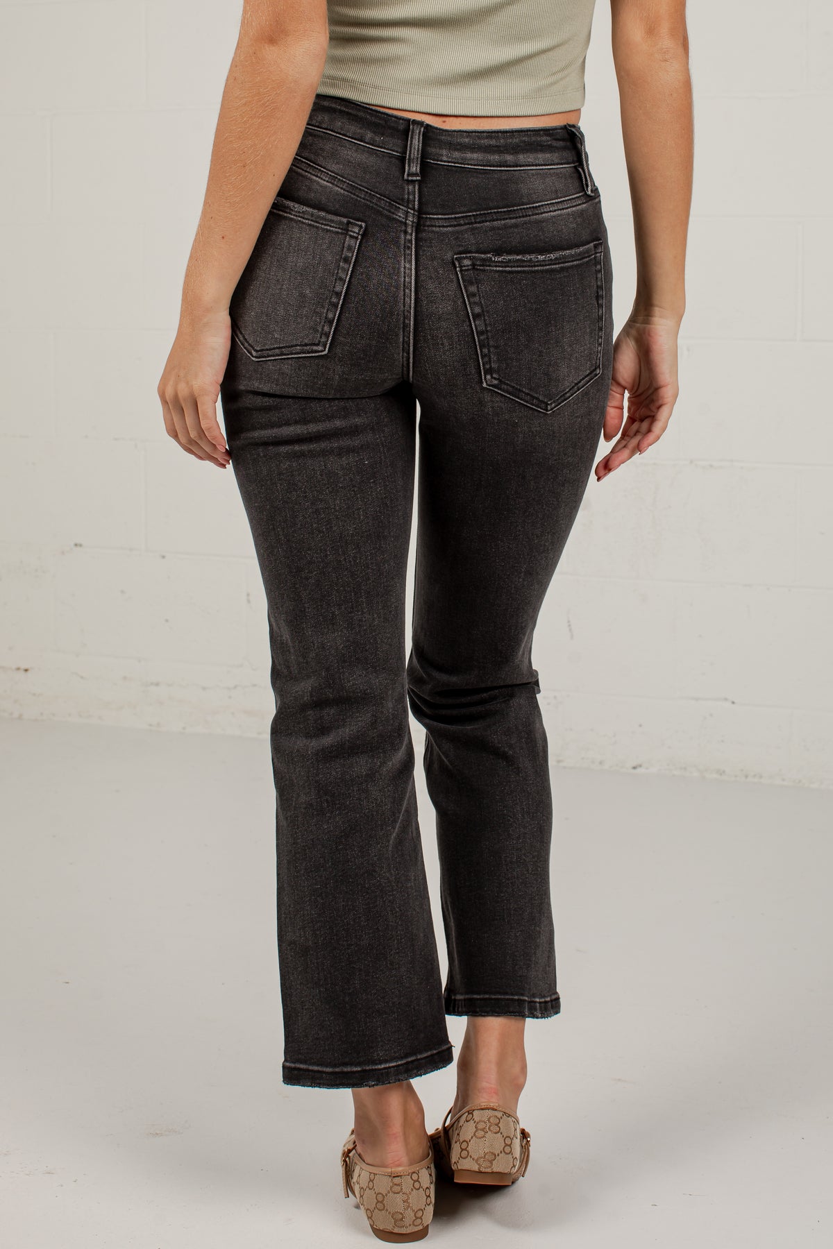 Rear view of a person wearing dark charcoal gray cropped flare jeans with a faded wash, paired with a light beige top and tan flat shoes, against a white brick wall background.