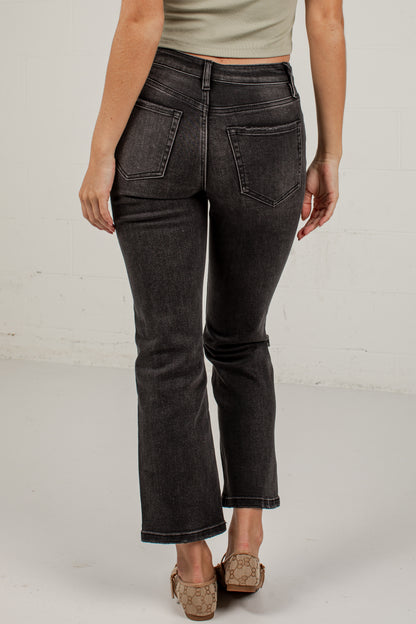 Rear view of a person wearing dark charcoal gray cropped flare jeans with a faded wash, paired with a light beige top and tan flat shoes, against a white brick wall background.