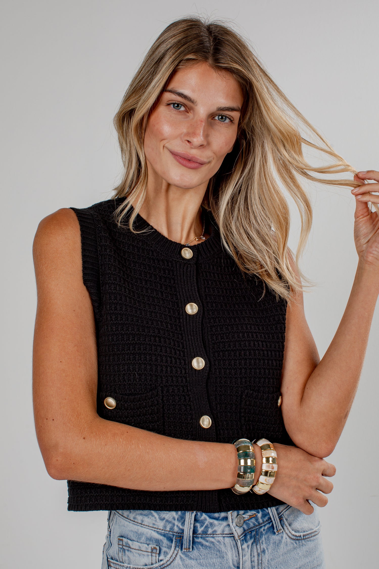A woman wearing a black sleeveless textured knit top with gold buttons, paired with light blue jeans, against a plain light gray studio background.