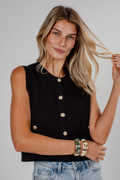 A woman wearing a black sleeveless textured knit top with gold buttons, paired with light blue jeans, against a plain light gray studio background.