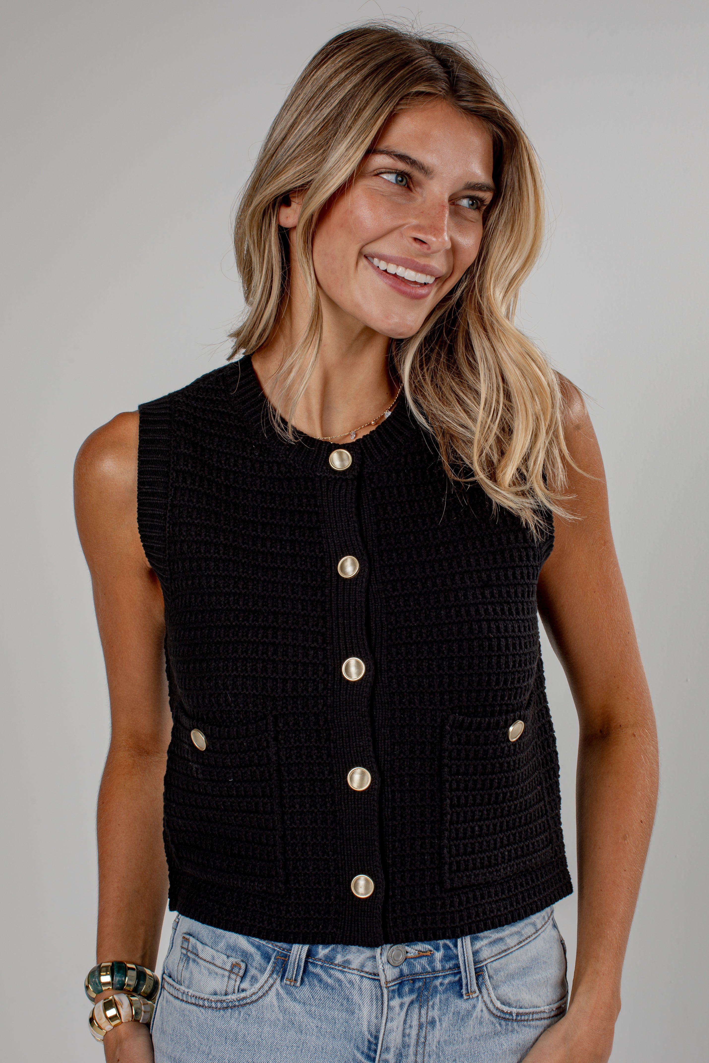 A woman wearing a sleeveless black textured knit vest with gold-tone buttons, paired with light-wash blue jeans, photographed against a neutral gray studio background.