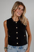 A woman wearing a sleeveless black textured knit vest with gold-tone buttons, paired with light-wash blue jeans, photographed against a neutral gray studio background.