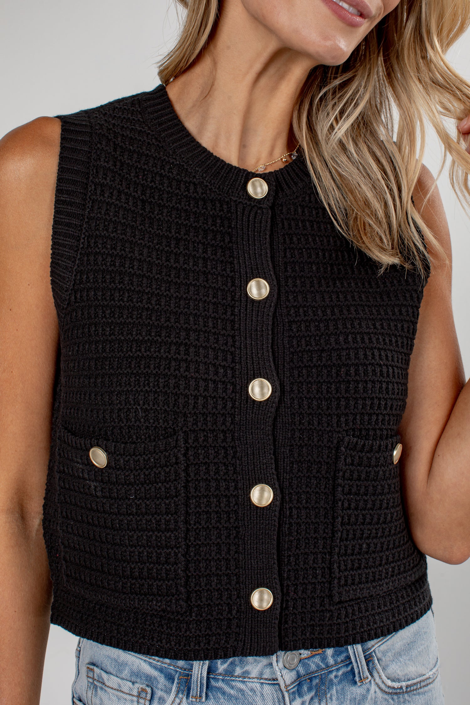 A close-up of a woman wearing a sleeveless black crocheted vest with gold-tone buttons down the front and two matching side pockets, paired with light blue jeans.