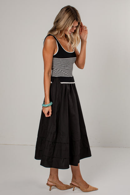 A woman modeling a sleeveless midi dress with a black and white horizontally striped bodice and a full black tiered skirt, paired with tan heeled sandals against a plain light gray studio background.