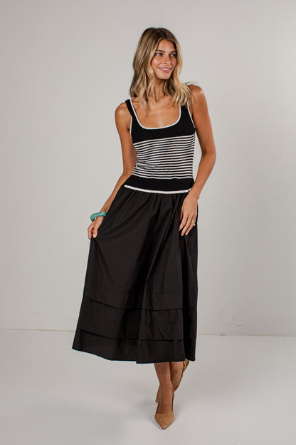 A woman modeling a sleeveless midi dress with a black and white horizontally striped bodice and a solid black tiered skirt, standing against a light gray studio background.