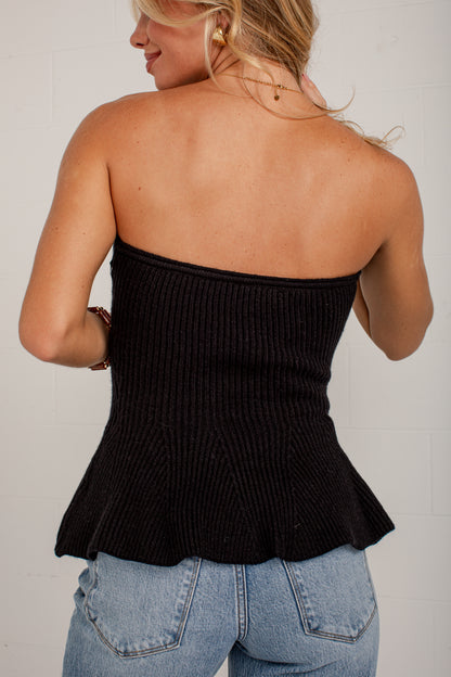 A woman shown from the back wearing a black ribbed strapless bandeau-style top with a peplum hem, paired with light blue jeans, against a white brick wall.