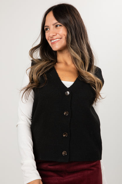 Person wearing a black sweater vest over a white long-sleeve shirt on a plain background