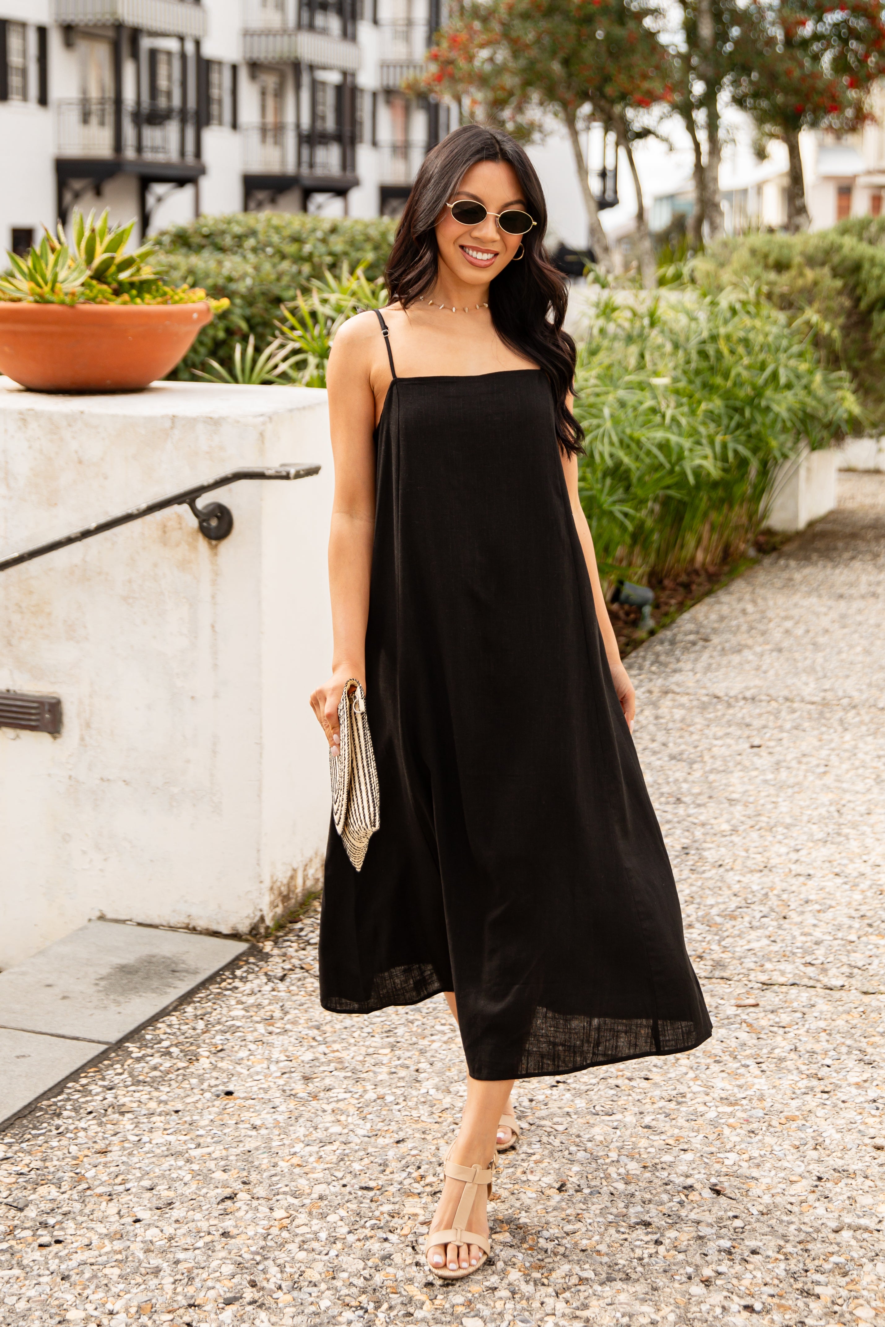 Woman in a black dress standing on a sidewalk with greenery and buildings in the background