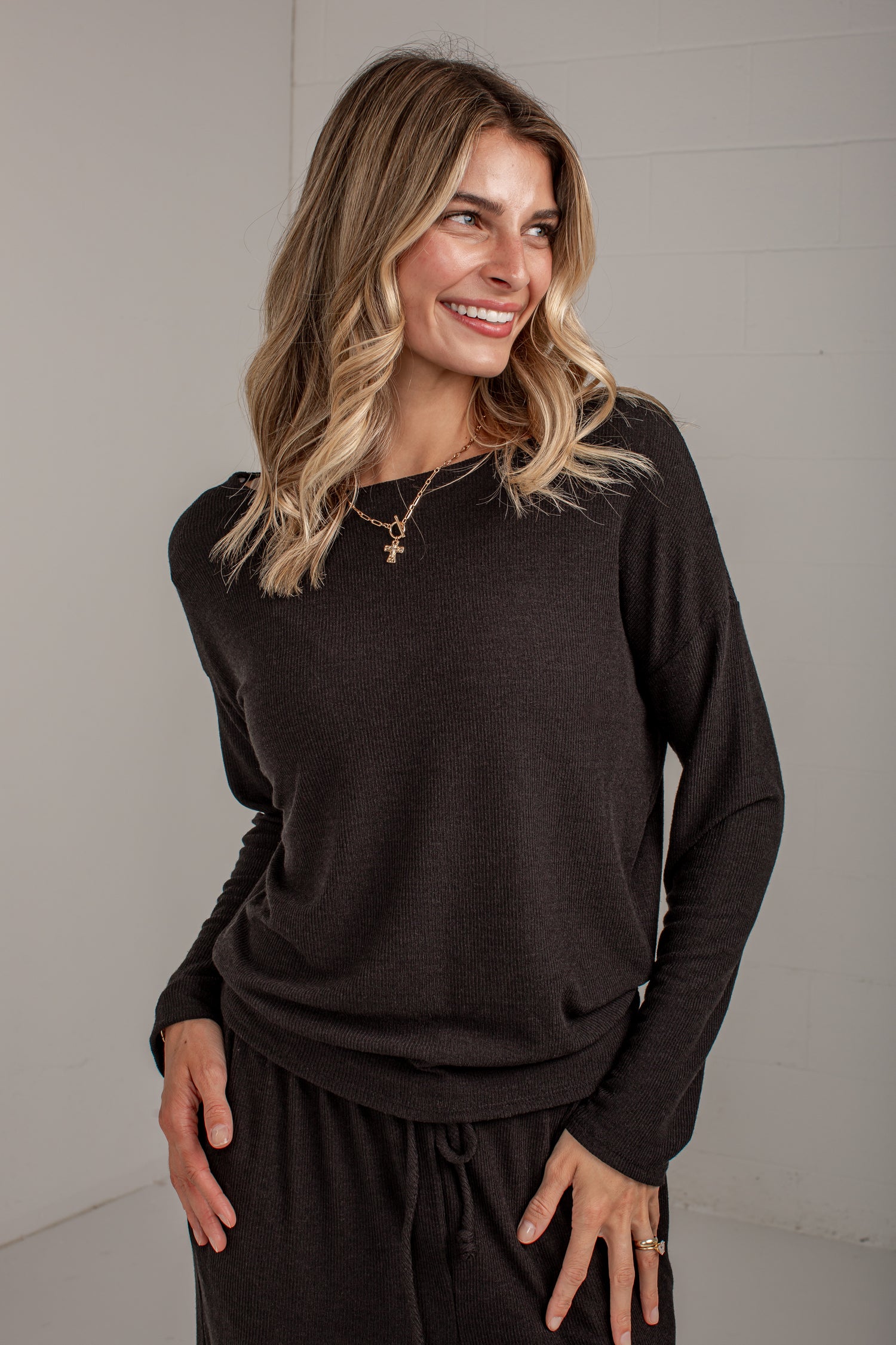 A woman wearing a dark brown long-sleeve knit top and matching dark pants, standing against a light gray wall.