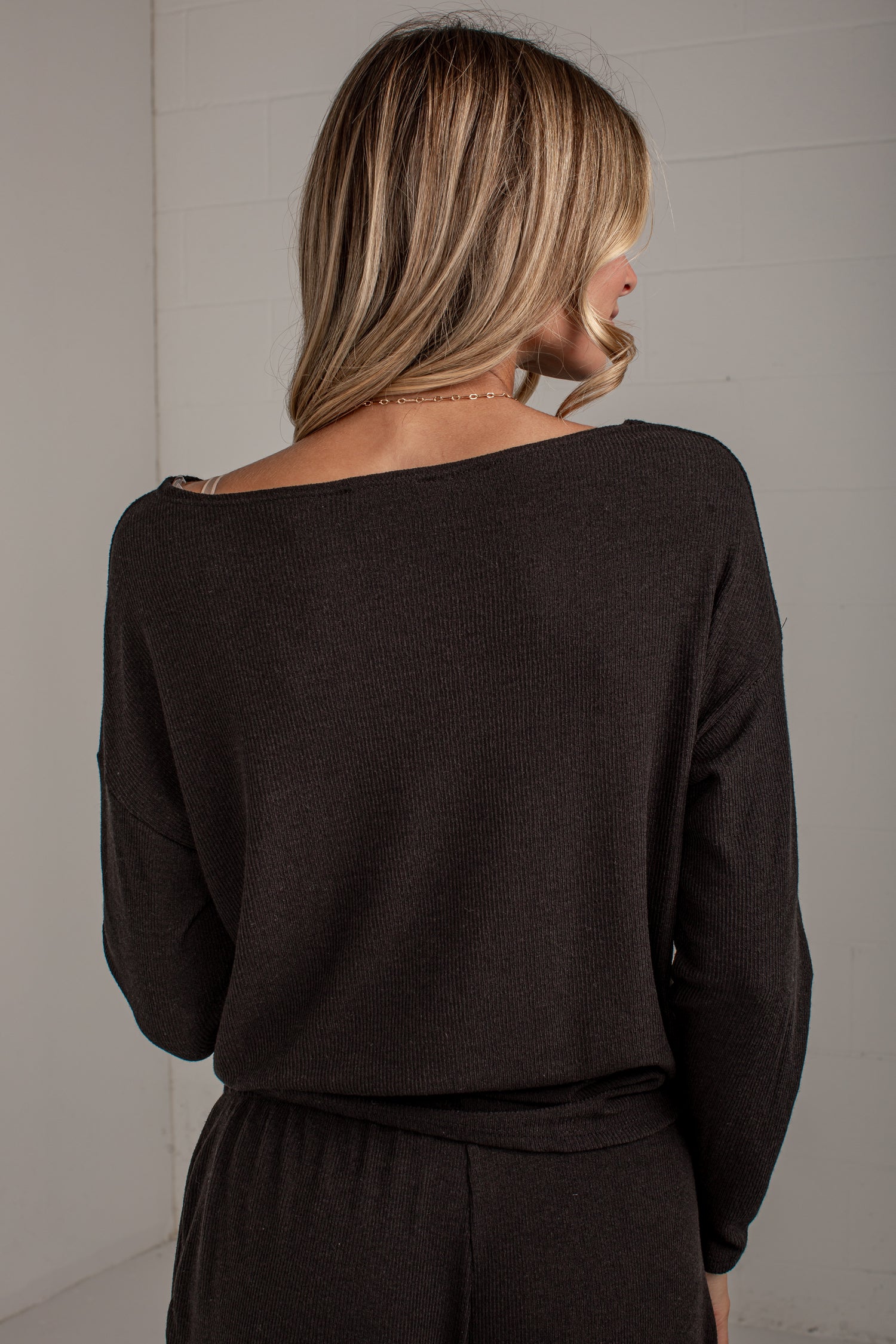 A woman viewed from behind wearing a dark brown off-the-shoulder knit sweater and matching dark bottoms, standing against a light gray paneled wall.