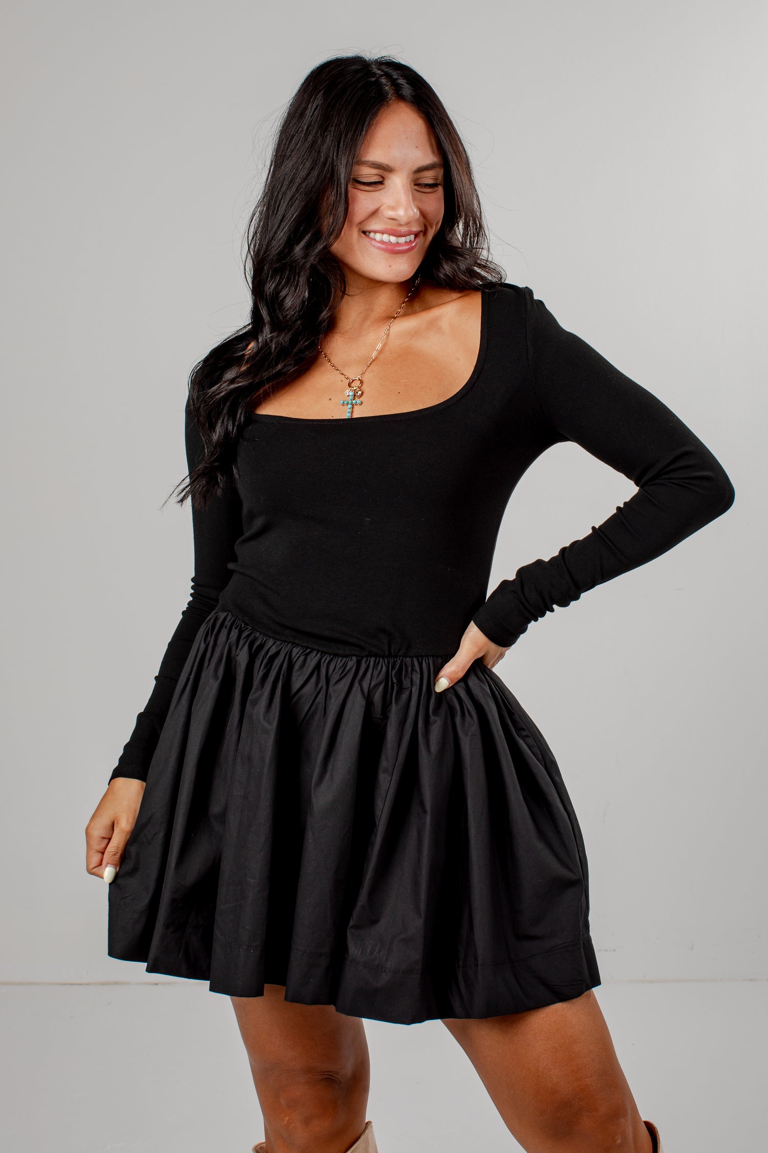 A woman modeling a black long-sleeve mini dress with a scoop neckline and a gathered flared skirt, posed against a light gray studio background.