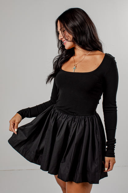 A woman modeling a black long-sleeve scoop-neck dress with a flared skirt, photographed against a plain light gray studio background.