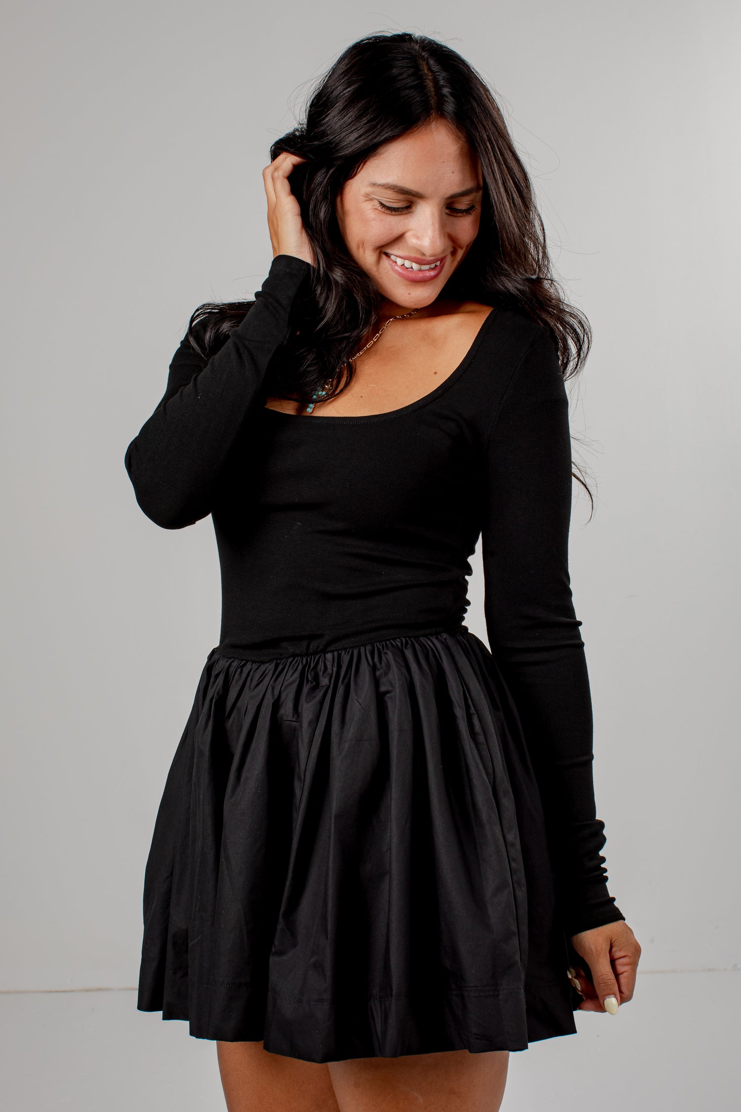 A woman wearing a black long-sleeve scoop-neck top and a black pleated mini skirt, posing against a light gray studio background.