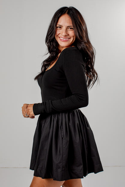 A woman modeling a black long-sleeve skater dress with a pleated skirt, photographed against a neutral light gray studio background.