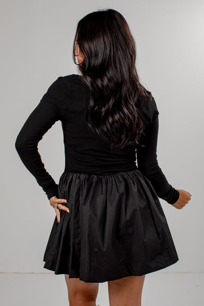 A woman shown from the back wearing a black long-sleeve top and a black pleated mini skirt, standing against a plain light gray studio background.