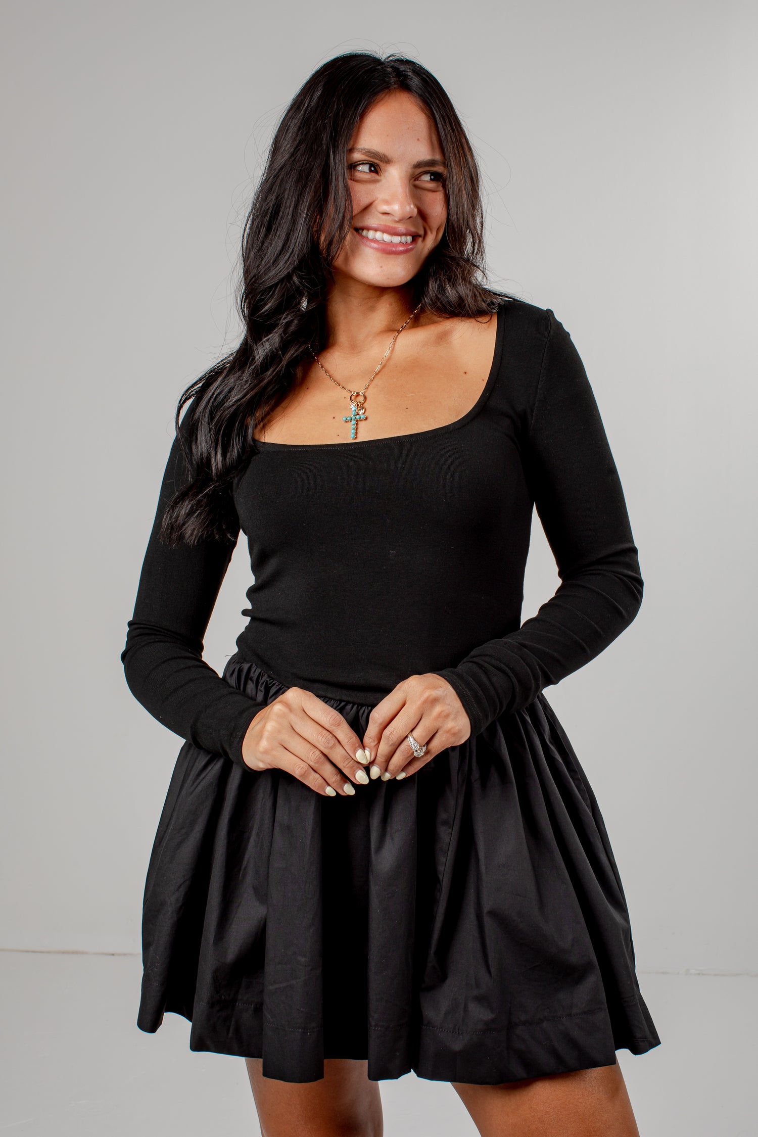 A woman modeling a black long-sleeve scoop-neck top with a matching black pleated mini skirt, photographed against a neutral light gray studio background.