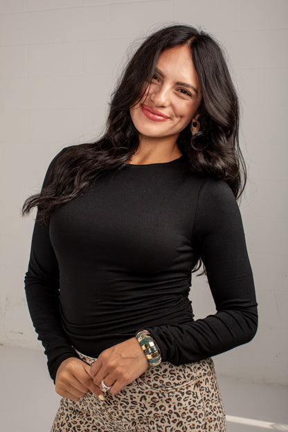 A woman with long dark hair wearing a fitted black long-sleeve top and a leopard print skirt, standing against a light gray wall.