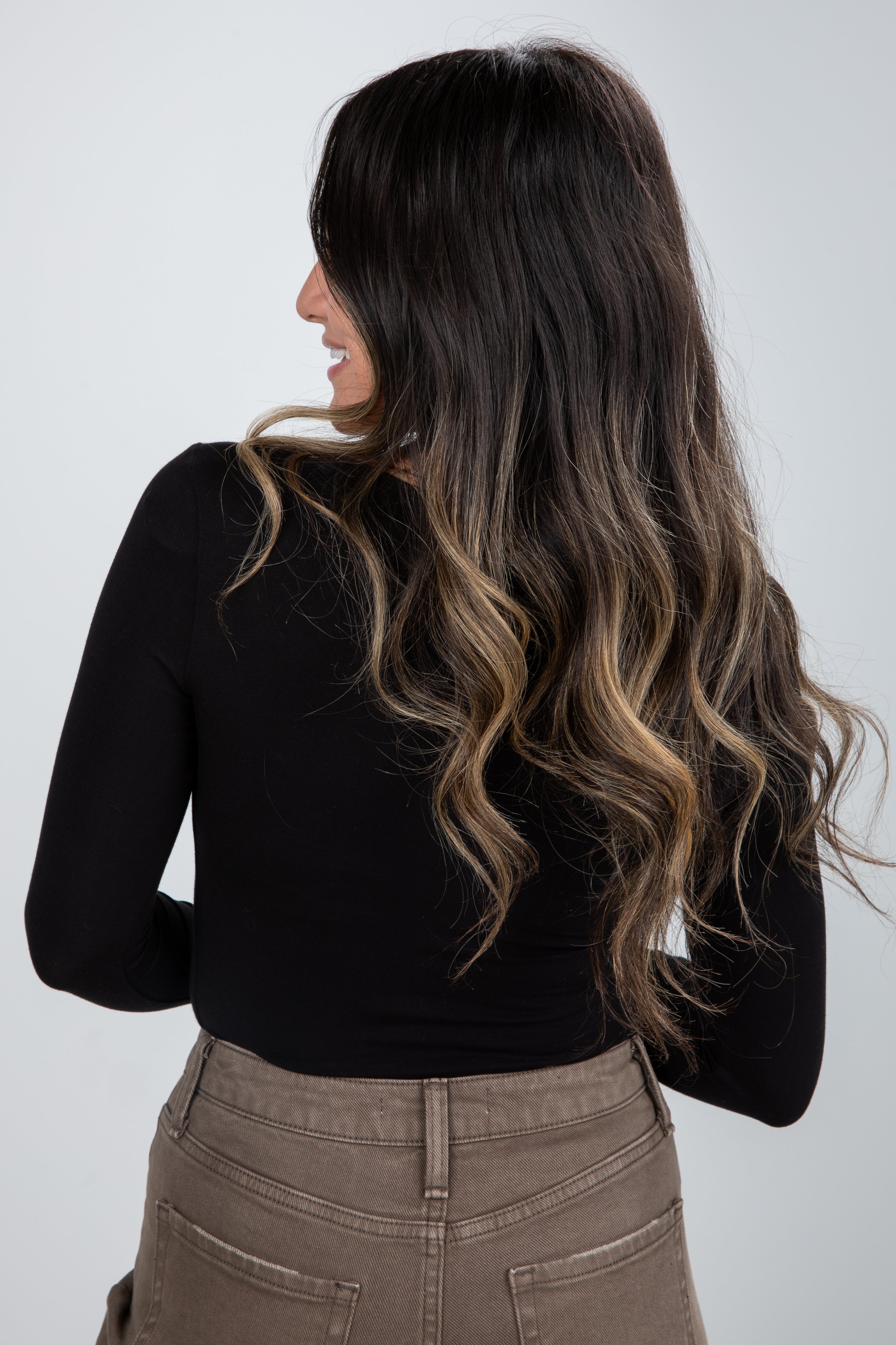 Woman with long, wavy hair wearing a black top and beige pants on a white background