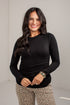A woman with long dark hair smiling, wearing a fitted black long-sleeve top and leopard print pants, standing against a light gray wall.