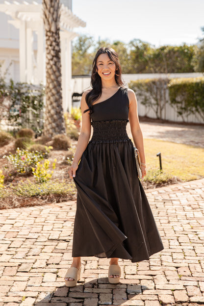 Woman in a black one-shoulder dress standing on a paved walkway with greenery in the background