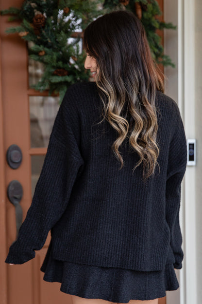 Person wearing a black sweater and skirt standing in front of a door with greenery.