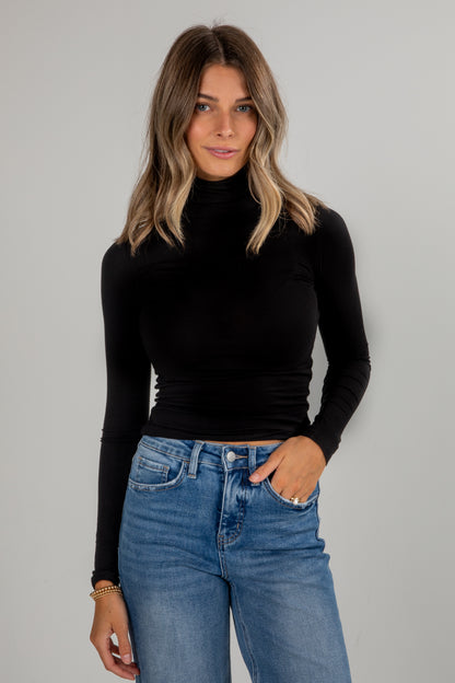 Woman wearing a black turtleneck and blue jeans against a gray background