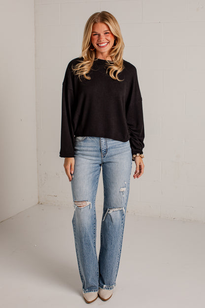 A woman modeling a black long-sleeve crewneck top and light-wash distressed jeans with ripped knees, standing against a white brick wall.