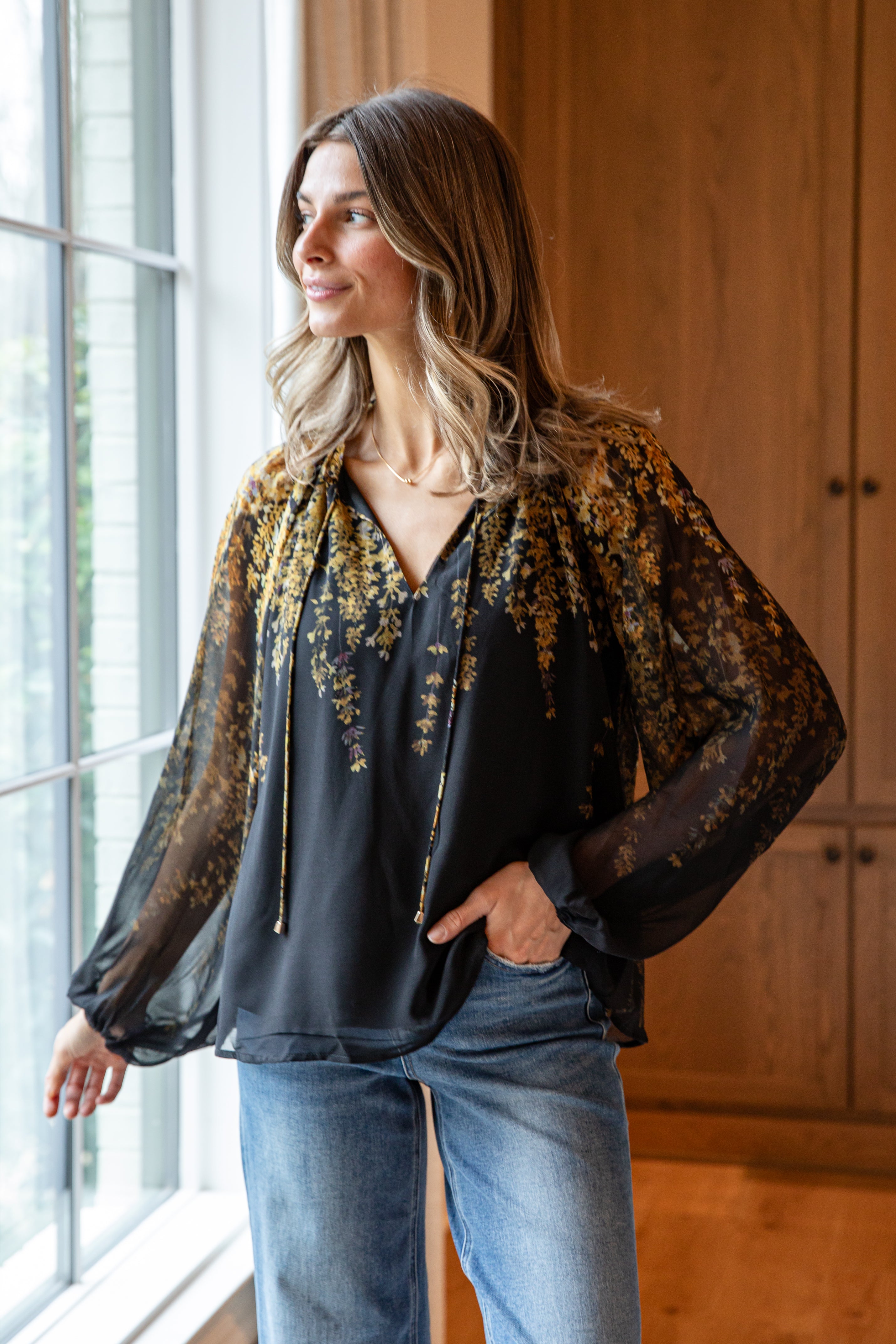 Woman wearing a black blouse with sheer sleeves and floral scarf, standing by a window.