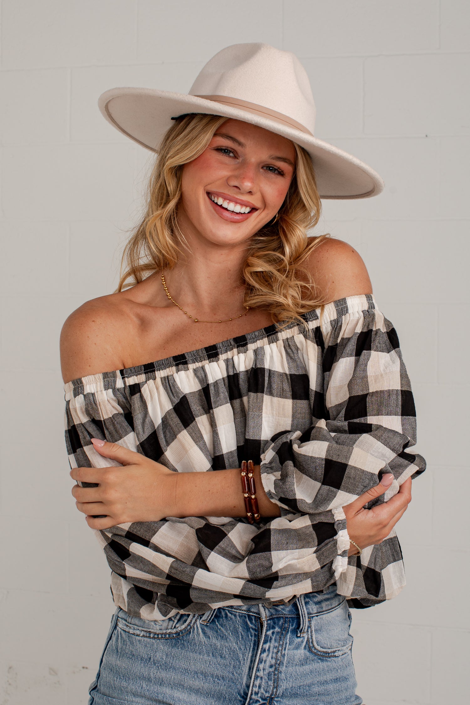 A smiling woman wearing an off-the-shoulder black and white buffalo plaid top, a cream wide-brim fedora hat, and blue jeans, posing against a light gray brick wall.