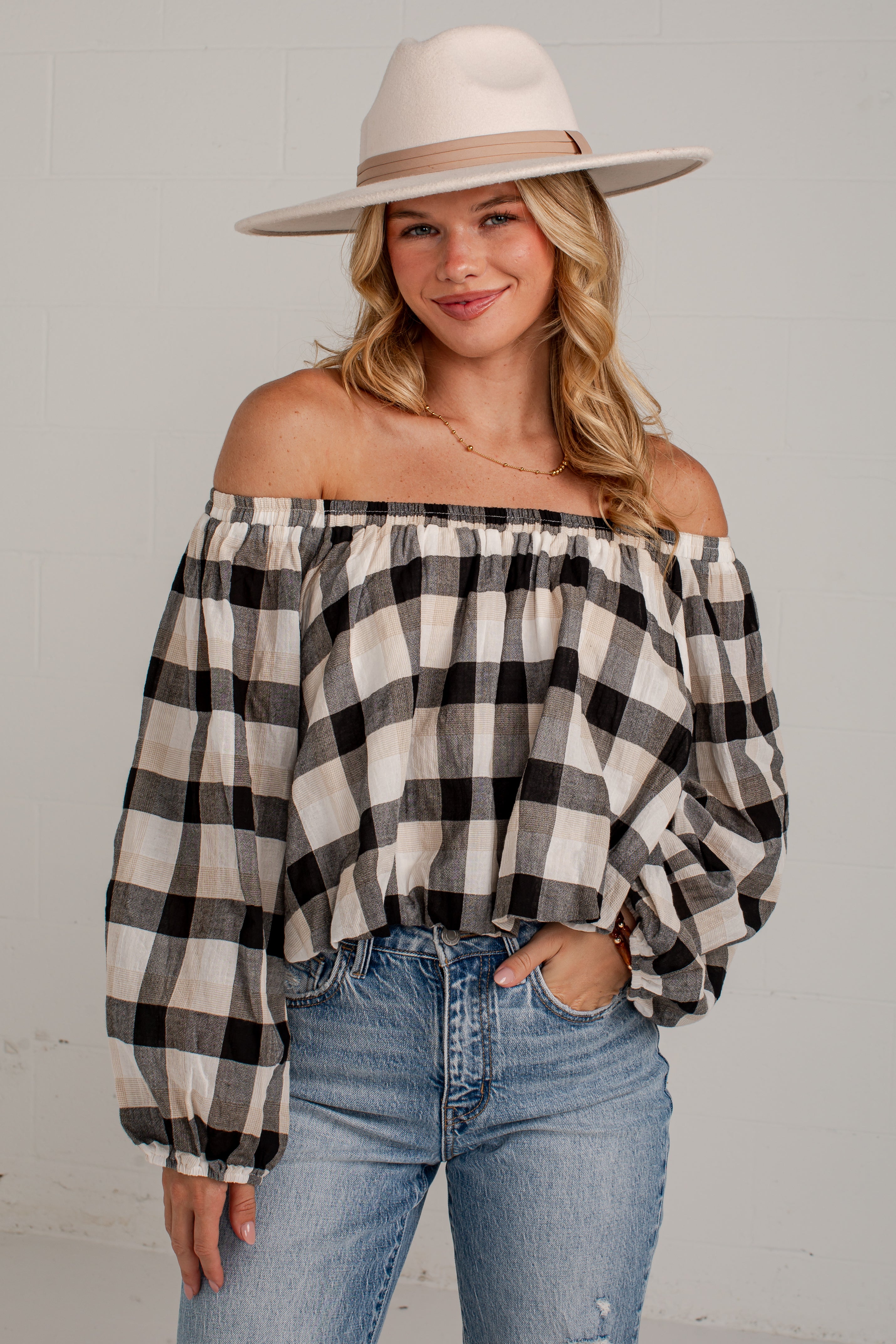 A woman wearing an off-the-shoulder black and white buffalo check blouse with voluminous long sleeves, paired with light-wash blue jeans and a beige wide-brim fedora hat, posing against a light gray brick wall.