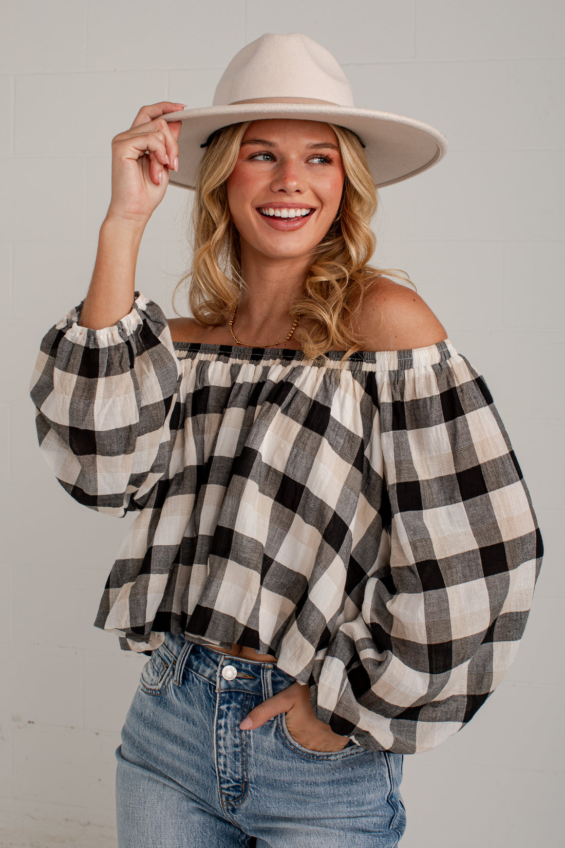 A woman wearing a black and white buffalo check off-the-shoulder blouse with voluminous sleeves, paired with blue jeans and a beige wide-brim hat, posing against a light gray brick wall.