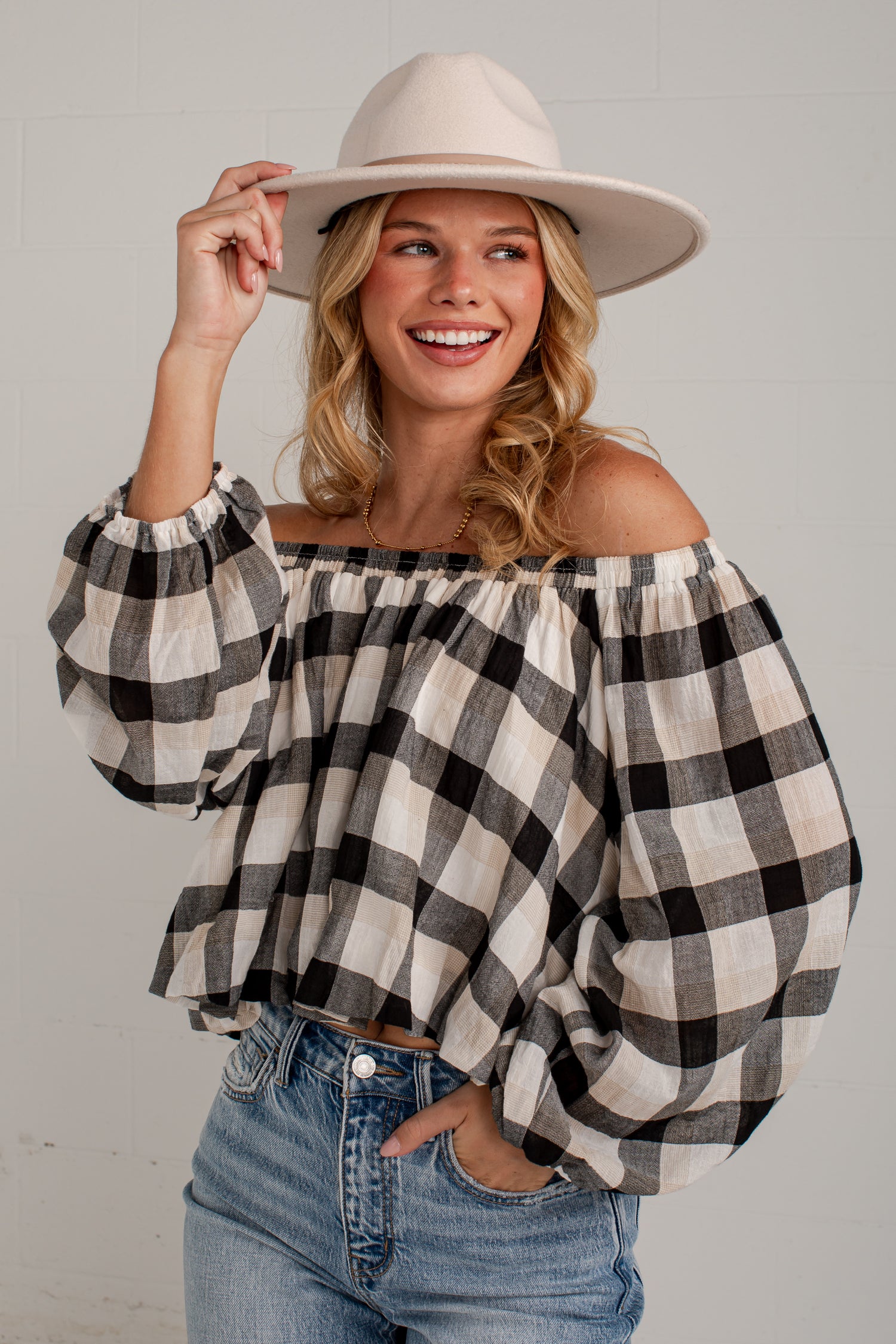 A woman wearing a black and white buffalo check off-the-shoulder blouse with voluminous sleeves, paired with blue jeans and a beige wide-brim hat, posing against a light gray brick wall.