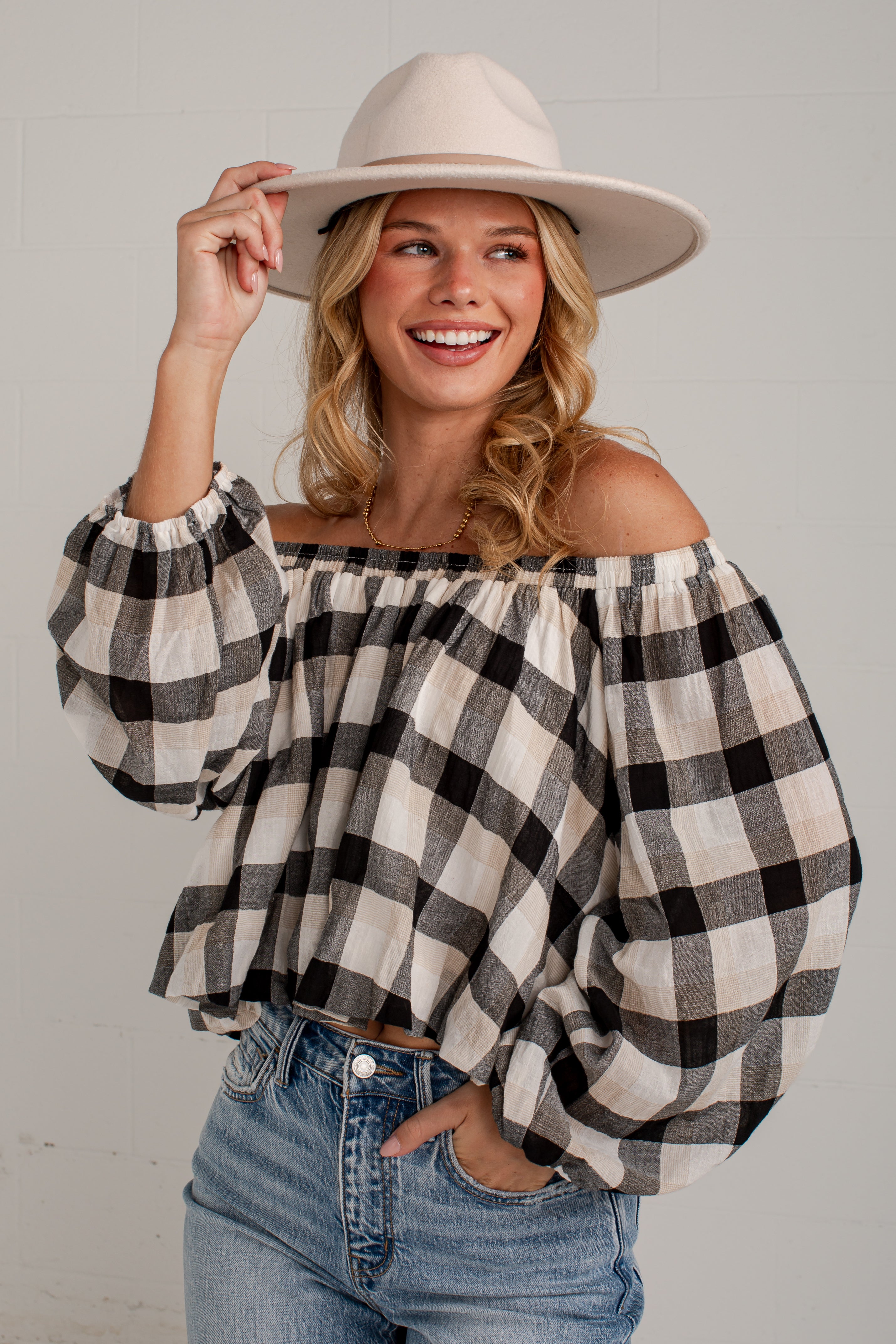 A woman wearing a black and white buffalo check off-the-shoulder blouse with voluminous sleeves, paired with blue jeans and a beige wide-brim hat, posing against a light gray brick wall.