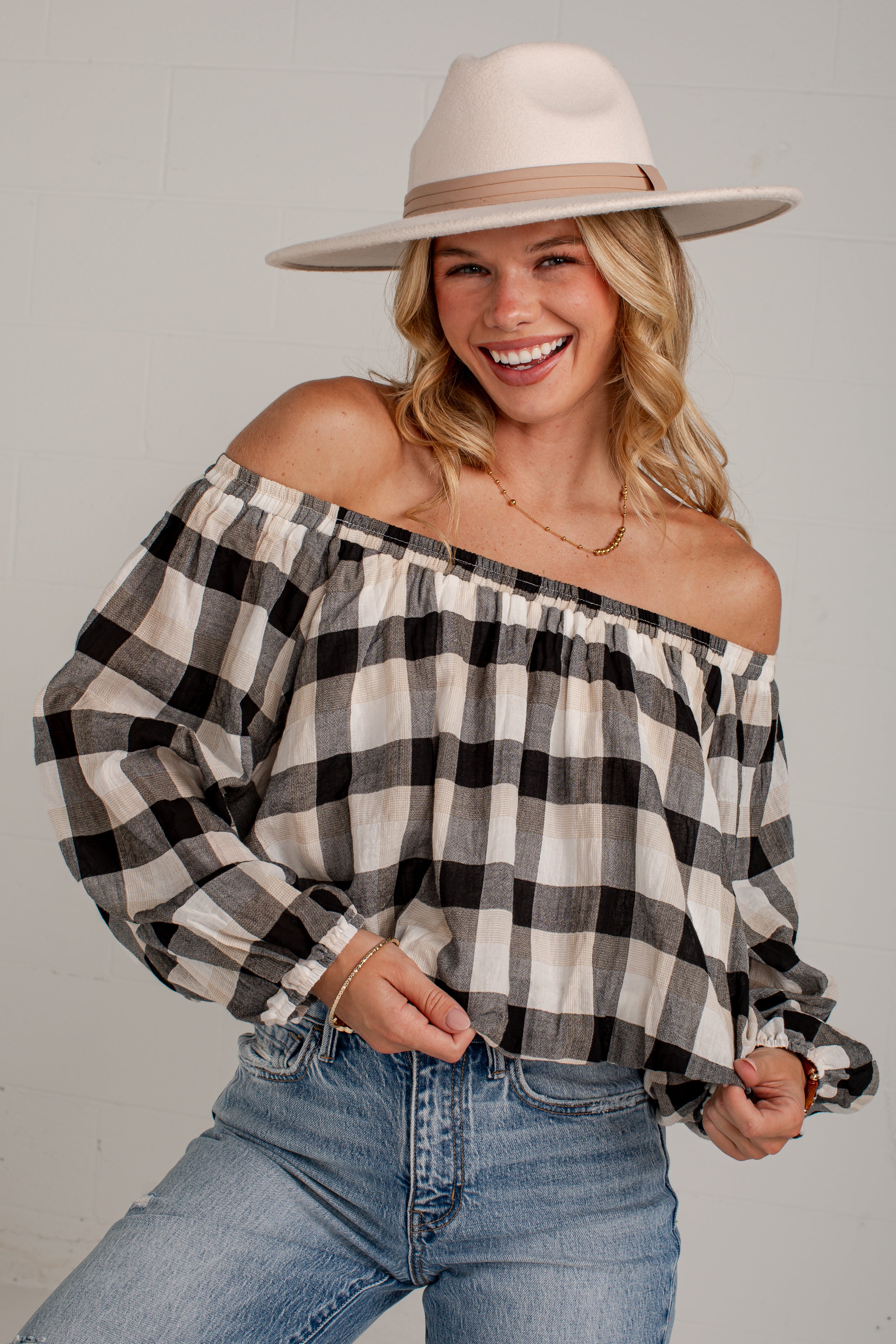A woman wearing a black, white, and gray plaid off-the-shoulder blouse with blue jeans and a beige wide-brim hat, posing against a light gray wall.