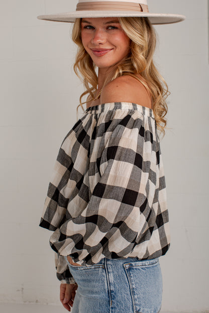 A woman wearing an off-the-shoulder black and beige buffalo plaid blouse with voluminous sleeves, paired with light blue jeans and a beige wide-brim hat, posing against a white wall.