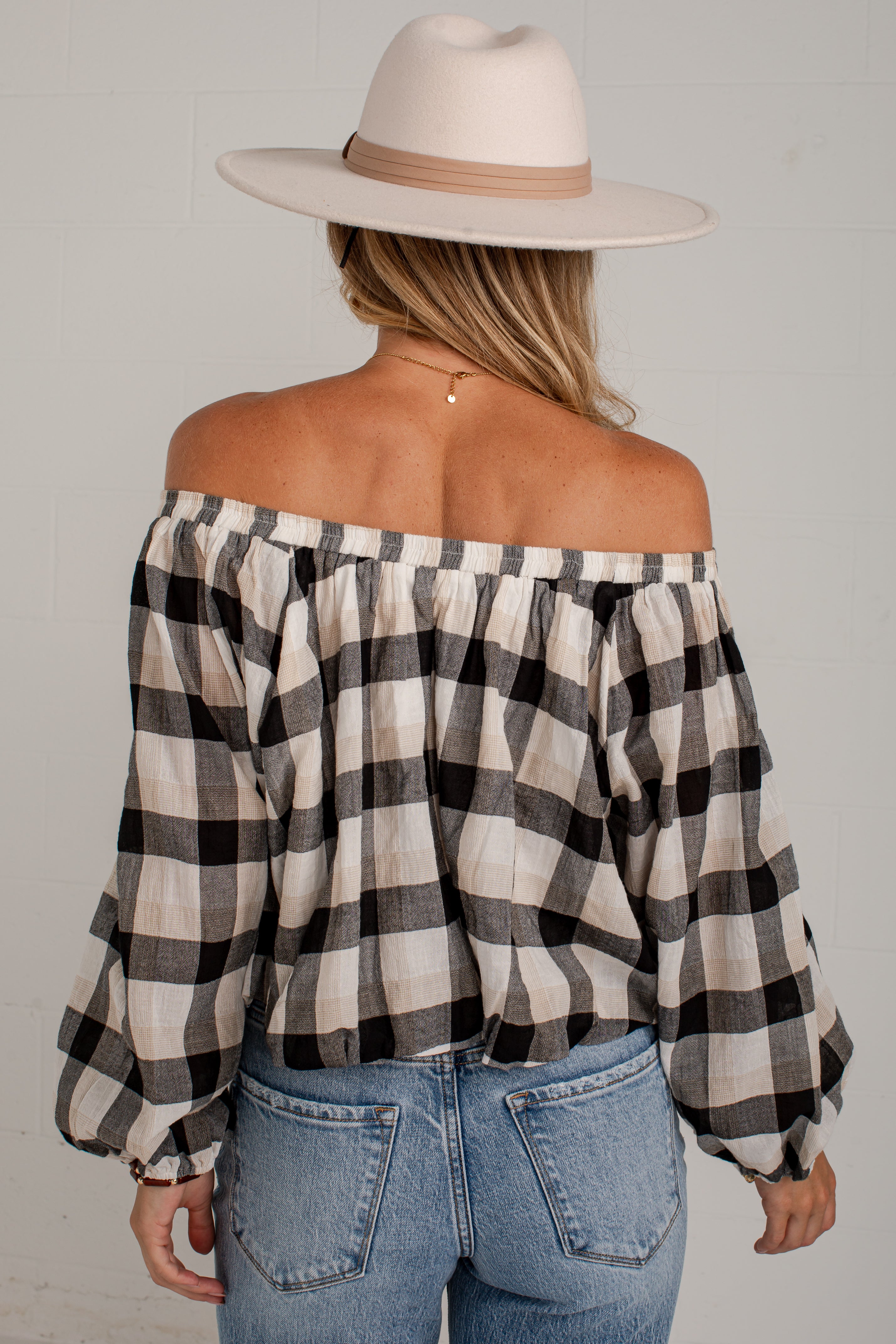 A woman shown from the back wearing an off-the-shoulder black and beige plaid blouse, blue jeans, and a cream wide-brim hat against a light gray wall.