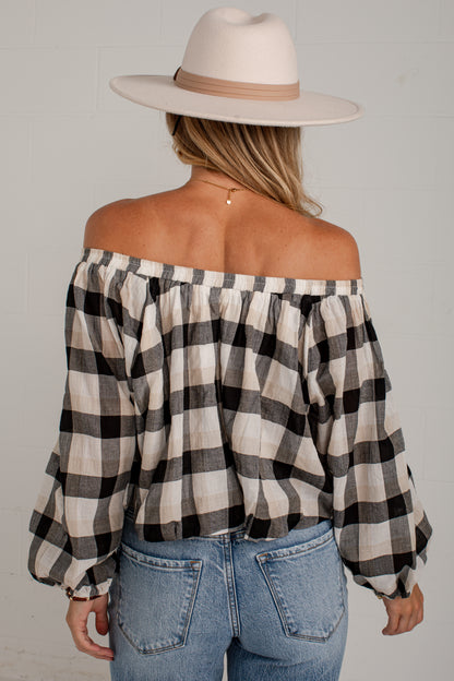 A woman shown from the back wearing an off-the-shoulder black and beige plaid blouse, blue jeans, and a cream wide-brim hat against a light gray wall.